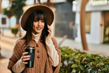 Kışlık şapkalı esmer kadın telefonda konuşuyor ve şehirde bir fincan kahve içiyor.