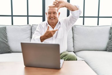 Sunbae evde dizüstü bilgisayar kullanıyor koltukta oturuyor gülümsüyor ve elleri ve parmakları gülen yüzlü bir çerçeve yapıyor. yaratıcılık ve fotoğrafçılık kavramı. 