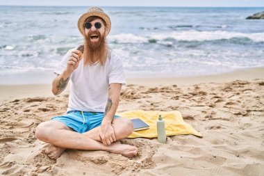 Genç kızıl adam dondurma yiyor kumsalda havlunun üzerinde oturuyor..