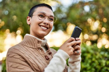 Kısa saçlı genç İspanyol kadın akıllı telefon kullanarak mutlu bir şekilde gülümsüyor.
