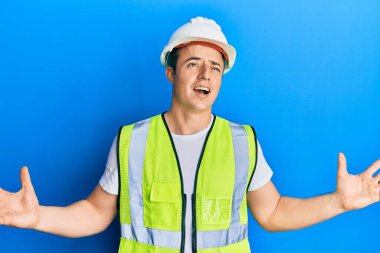 Güvenlik kaskı ve yansıtıcı ceket giyen yakışıklı genç adam deli gibi bağırıyor ve agresif bir ifadeyle bağırıyor ve kollarını kaldırıyor. hüsran kavramı. 