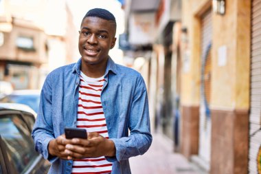 Genç Afrikalı Amerikalı adam şehirde akıllı telefon kullanarak mutlu bir şekilde gülümsüyor..