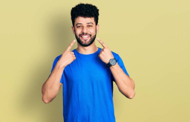 Sakallı, sıradan mavi tişörtlü, ağzı açık gülümseyen, parmakları işaret eden ve neşeli gülümsemeye zorlayan genç bir Arap. 