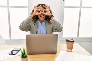 Güzel bir İspanyol iş kadını ofisinde oturmuş dizüstü bilgisayarla çalışıyor. Dürbün gibi jest yapıyor. Gözleri parmaklarından bakıyor. çılgın ifade. 