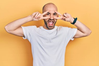 Gündelik beyaz tişört giyen genç kel adam barış sembolü yapıyor. Yüzünde parmaklar, gülümseyerek zaferi gösteriyor. 