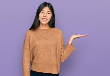 Güzel, genç Asyalı kadın, günlük kışlık süveter giyer, gülümser, mutlu bir şekilde sunar ve avuç içi kameraya bakar.. 