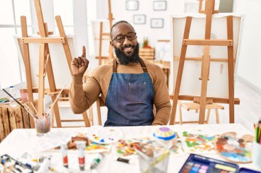 African american artist man at art studio showing and pointing up with finger number one while smiling confident and happy. 
