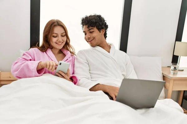 Młoda Para Uśmiechnięta Szczęśliwa Korzystając Laptopa Smartfona Sypialni — Zdjęcie stockowe