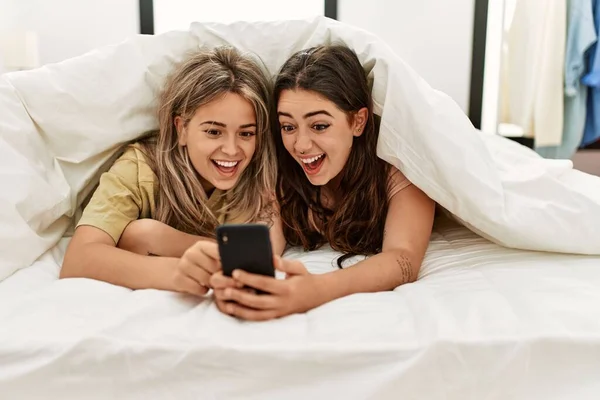 ベッドの上に横になっているシートでカバースマートフォンを使用して若いカップル — ストック写真