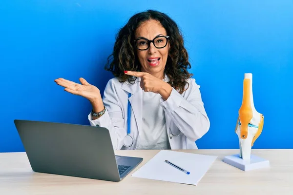 Beautiful middle age woman doctor at orthopedic clinic amazed and smiling to the camera while presenting with hand and pointing with finger.