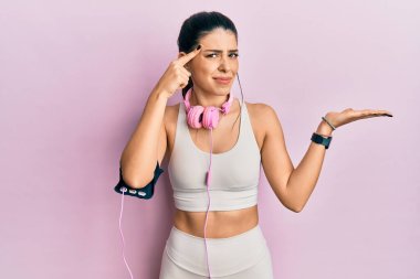 Spor kıyafetleri giyen ve kulaklık takan genç İspanyol bir kadın. Avuç içini açıp fotokopi alanını göstermekten rahatsız oluyor ve alnını işaret ediyor. Bunu bir düşün.. 