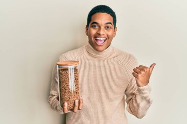 Sağlıklı tahıl gevreği dolu kavanozu tutan genç, yakışıklı İspanyol adam başparmağını yana uzatıyor ve ağzı açık gülümsüyor. 