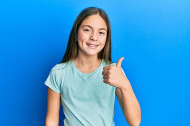 Güzel, esmer, beyaz tişört giyen küçük bir kız mutlu bir el hareketi yapıyor. Kameraya bakıp başarıyı gösteren ifade. 