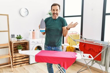 Sakallı orta yaşlı bir adam evde kıyafetlerini ütülüyor başarıyı mutlu bir gülümsemeyle kutluyor ve kazanan yüz ifadesi kaldırılmış bir el ile. 