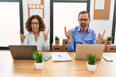 Orta yaşlı İspanyol kadın ve erkek ofiste dizüstü bilgisayarla oturup çılgın bir ifadeyle bağırarak elleri havada rock sembolü yapıyorlar. Müzik yıldızı. ağır kavram. 