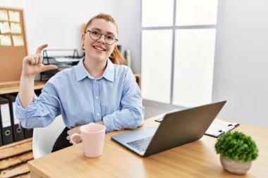 Ofiste çalışan genç kızıl saçlı kadın bilgisayarlı dizüstü bilgisayar kullanıyor gülümsüyor ve el hareketleriyle hareket ediyor parmaklarıyla işaret yapıyor ve kameraya bakıyor. ölçüm kavramı. 