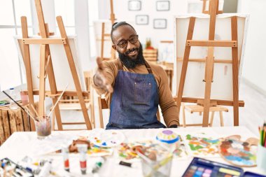 African american artist man at art studio smiling friendly offering handshake as greeting and welcoming. successful business. 
