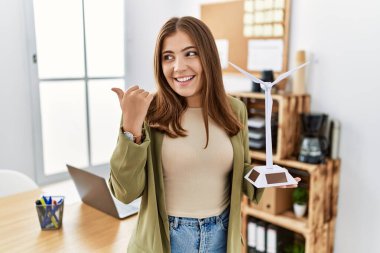 Genç esmer kadın ofisinde yenilenebilir elektrik için güneş değirmenini tutuyor başparmağını yana uzatıyor ve ağzı açık gülümsüyor. 