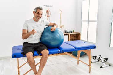 Orta yaşlı İspanyol adam ağrı kesici kliniğinde elinde pilates topuyla seni ve kamerayı işaret ediyor. 