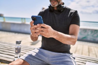 Orta yaşlı İspanyol adam deniz kenarında akıllı telefon kullanarak spor giyer.
