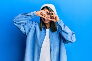 Hoş yün şapkalı genç Latin kadın kalp şeklinde el ve parmaklarıyla tabelanın içinden bakıp gülümsüyor. 