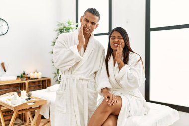 Young latin couple wearing towel standing at beauty center touching mouth with hand with painful expression because of toothache or dental illness on teeth. dentist concept. 