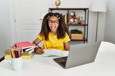 Young african american girl doing homework at home sticking tongue out happy with funny expression. emotion concept. 