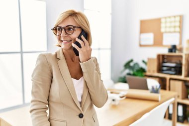 Orta yaşlı sarışın kadın, mimarlık stüdyosunda çalışan akıllı telefondan konuşuyor.
