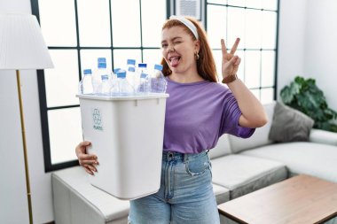 Geri dönüşüm sepetini elinde tutan, elinde plastik şişelerle kameraya gülümseyen ve parmaklarıyla zafer işareti yapan genç kızıl saçlı kadın. İki numara.. 