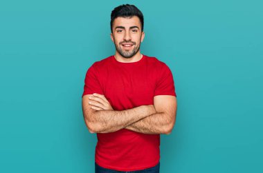 Kırmızı tişörtlü İspanyol sakallı, mutlu yüzlü, kollarını kavuşturup kameraya bakan adam. pozitif kişi. 