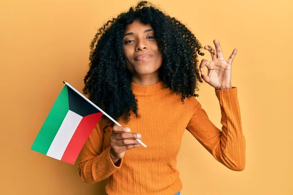Afro saçlı, elinde kuveyt bayrağı olan Afro-Amerikalı bir kadın parmaklarıyla tamam işareti yapıyor, gülümseyerek mükemmel bir sembol oluşturuyor. 