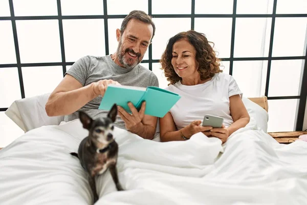 中年时 他的惊慌失措伴侣读书和使用智能手机 在家和狗躺在床上 — 图库照片