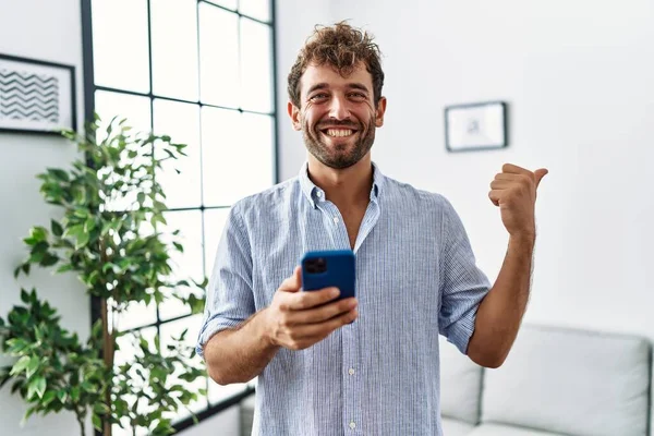 Jovem Homem Bonito Usando Smartphone Casa Apontando Polegar Para Lado — Fotografia de Stock