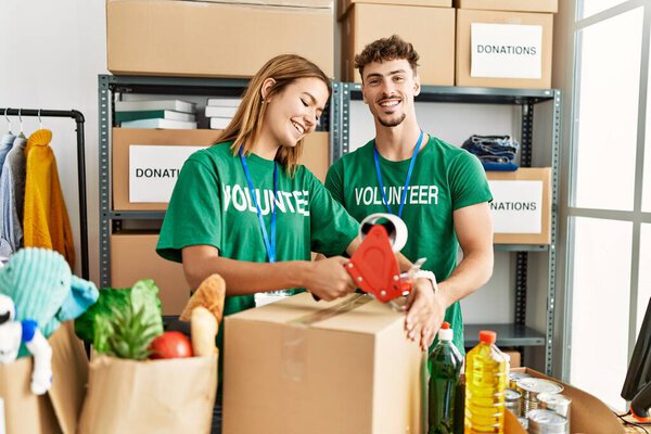 Young hispanic volunteer couple closing box with packing tape at charity center.