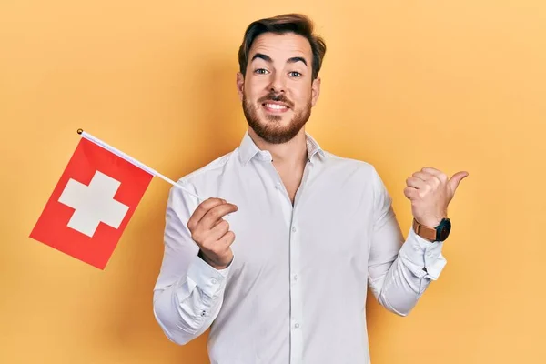 髭を生やしているハンサムな白人男性スイスの国旗を持ちます親指を指します側に笑みを浮かべて幸せなオープン口 — ストック写真