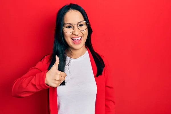 Beautiful hispanic woman with nose piercing wearing casual look and glasses doing happy thumbs up gesture with hand. approving expression looking at the camera showing success.