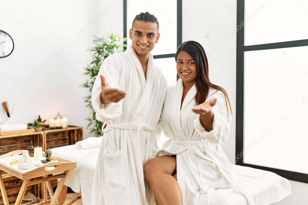 Young latin couple wearing towel standing at beauty center smiling friendly offering handshake as greeting and welcoming. successful business. 