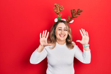 Orta yaşlı beyaz kadın şirin Noel geyiği boynuzları takıyor. Onuncu parmaklarını gösterip gösterirken kendinden emin ve mutlu bir şekilde gülümsüyor.. 