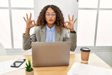 Güzel bir İspanyol iş kadını ofisinde oturmuş laptopla çalışıyor gözleri kapalı gülümsüyor parmaklarıyla meditasyon hareketi yapıyor. yoga konsepti. 