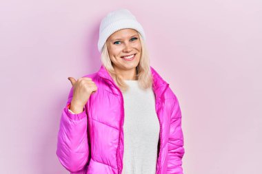 Güzel, beyaz, sarışın, yün şapkalı, kışlık ceketli, gülen yüzlü ve baş parmağıyla yüzünü gösteren bir kadın.. 