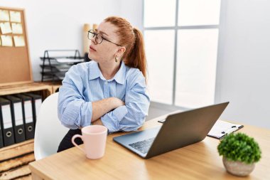 Ofiste çalışan genç, kızıl saçlı bir kadın bilgisayar kullanıyor. Kollarını kavuşturmuş ve kendinden emin bir şekilde yan tarafa bakıyor. 