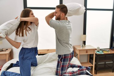 Genç beyaz çift mutlu bir şekilde gülümsüyor. Evdeki yatakta yastıklarla kavga ediyorlar..