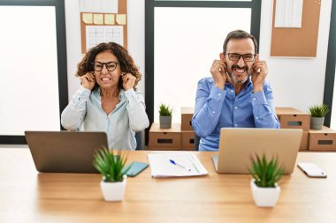 Orta yaşlı İspanyol kadın ve erkek ofiste dizüstü bilgisayarla oturup yüksek sesli müzik sesi için kulaklarını parmaklarıyla kapatıyor. sağır kavramı. 