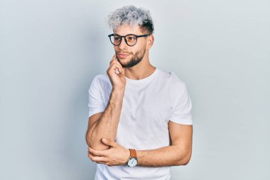 Modern boyalı saçlı, beyaz tişörtlü, eli çenesinde gözlüklü soru soran genç İspanyol bir adam. Düşünceli bir ifade. Gülümseyen ve düşünceli bir yüz. şüphe kavramı. 