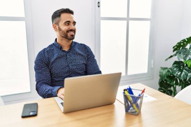 Ofiste sakallı, laptopla çalışan, yüzünde gülümseme ve doğal ifadeyle yan yana bakan genç bir İspanyol. Kendinden emin gülen. 