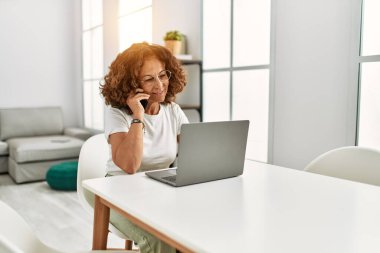 Orta yaşlı İspanyol kadın evde akıllı telefondan dizüstü bilgisayar kullanıyor.