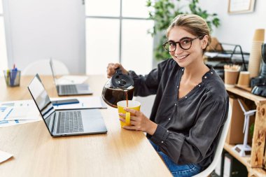 Kahve içen genç beyaz kadın ofiste çalışıyor.