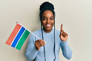 Saçları örgülü, gambiya bayrağı tutan, gülümseyen, mutlu yüzüyle işaret eden Afrikalı Amerikalı bir kadın. 