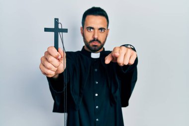 Sakallı, Katolik haçı tutan yakışıklı bir papaz kamerayı işaret ediyor ve kendine güveni tam bir jest yapıyor. 