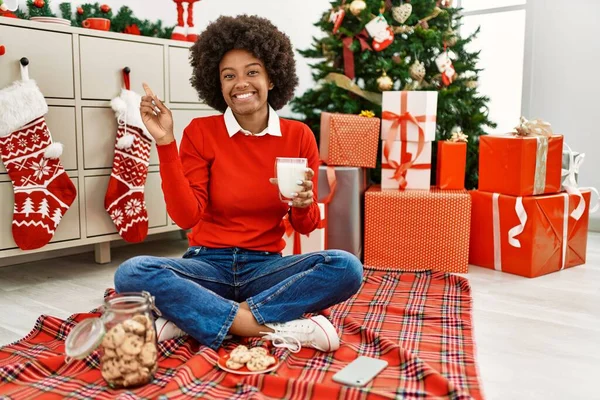 若いですアフリカ系アメリカ人女性とともにアフロ髪食べるペストリーによって座ってクリスマスツリー笑顔幸せなポインティングで手と指で側面 — ストック写真